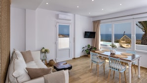 A modern living room with a sofa, a dining table with chairs, a TV, and a view of the sea through large windows. - Pure Seaside Calm, Far from Santorini’s Crowds (Thira)