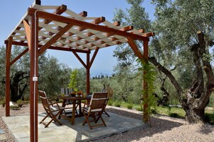 Una pérgola de madera con una mesa y sillas, rodeada de olivos y un cielo despejado.