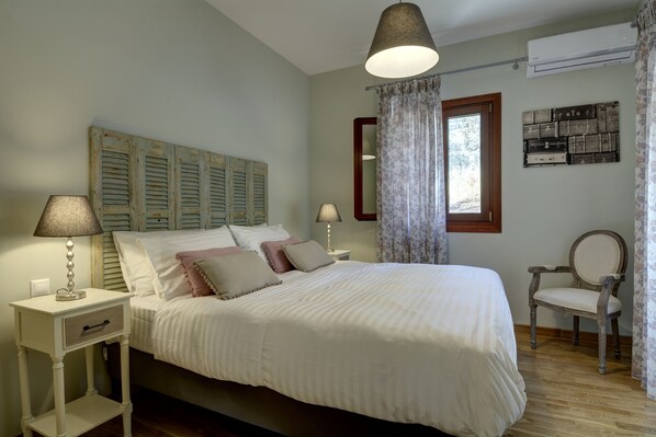 Una cama bien hecha con ropa blanca, dos lámparas de noche, un cabecero de madera, una silla y un aire acondicionado de pared.