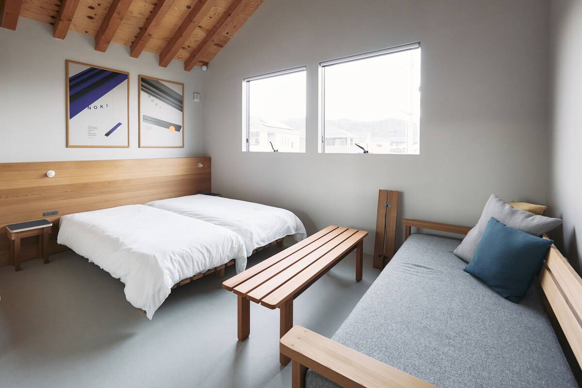 Quarto com duas camas, um banquinho, uma mesa pequena e janelas amplas.