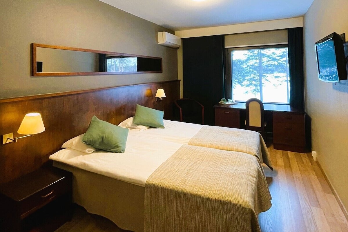A hotel room with a large bed, a bedside table, a lamp, a TV, and a window with a view of trees.