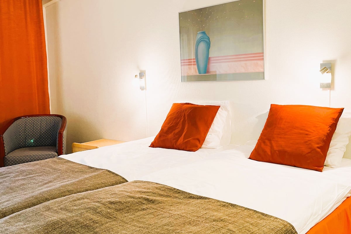 A hotel room with a bed, two pillows, a chair, an air conditioner, and a painting on the wall.