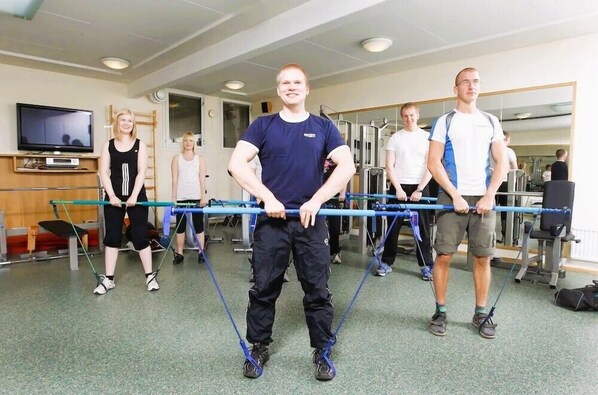 A fitness class in a gym with exercise equipment and a television in the background.