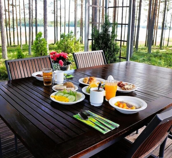 A wooden table set with breakfast items including croissants, eggs, and fruit, with a view of trees and a body of water in the background. - Herttua Hotel and Spa (Savonlinna)