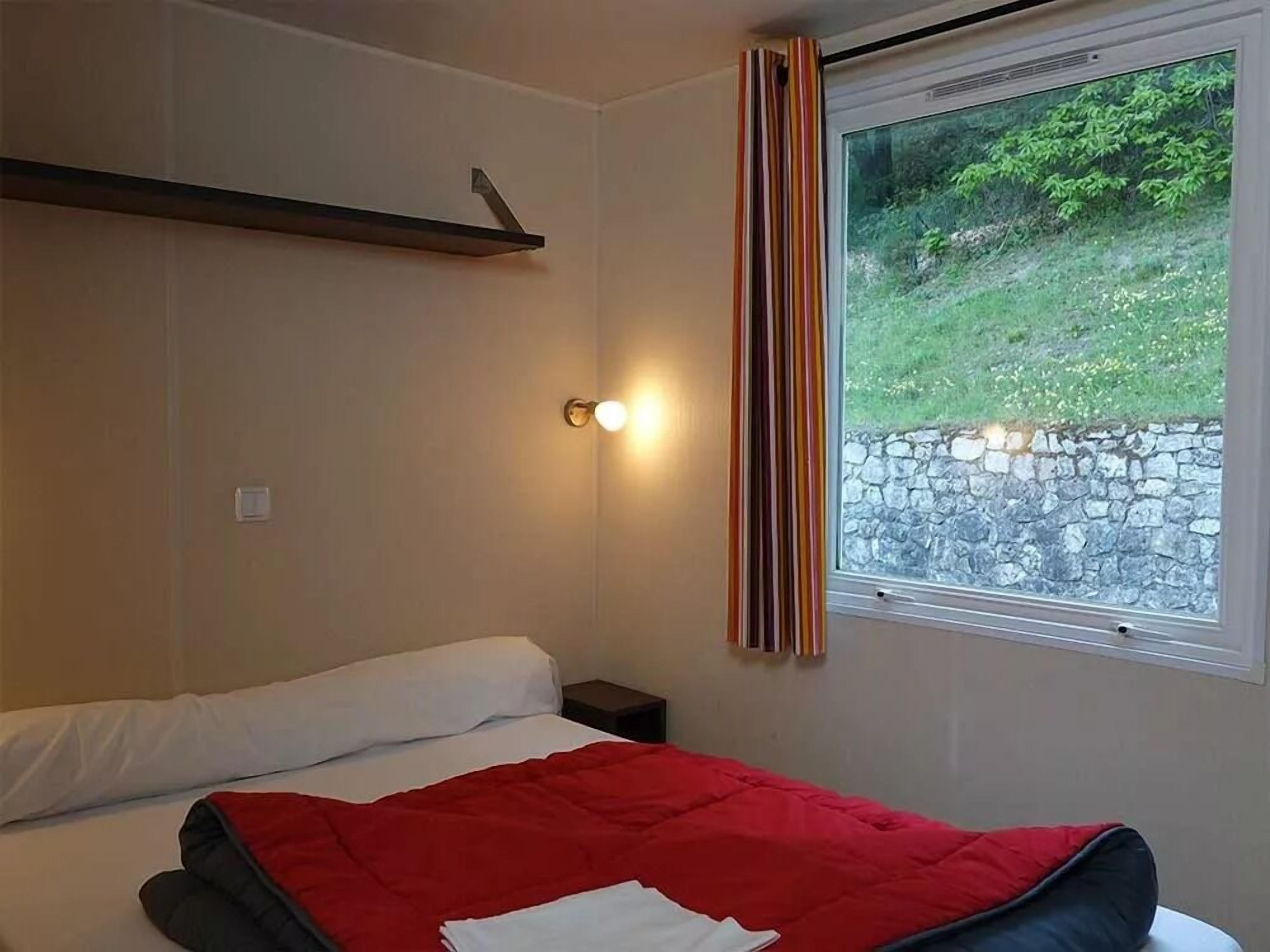 A bedroom with a bed, a bedside table, a wall-mounted shelf, a window with curtains, and a view of greenery outside.