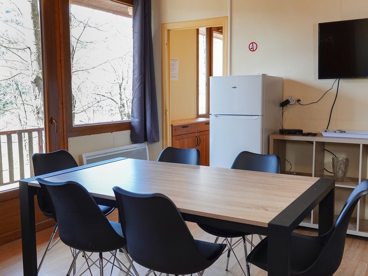 Una habitación con una mesa de madera y sillas negras, un refrigerador blanco, un televisor montado en la pared y una ventana con vista a árboles.