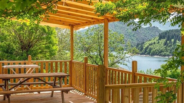 A wooden deck with picnic tables and chairs, overlooking a lake and forested mountains. - Camping Lac de Villefort (Pourcharesses)