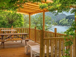 Una terraza de madera con mesas y sillas de picnic, con vistas a un lago y montañas boscosas.