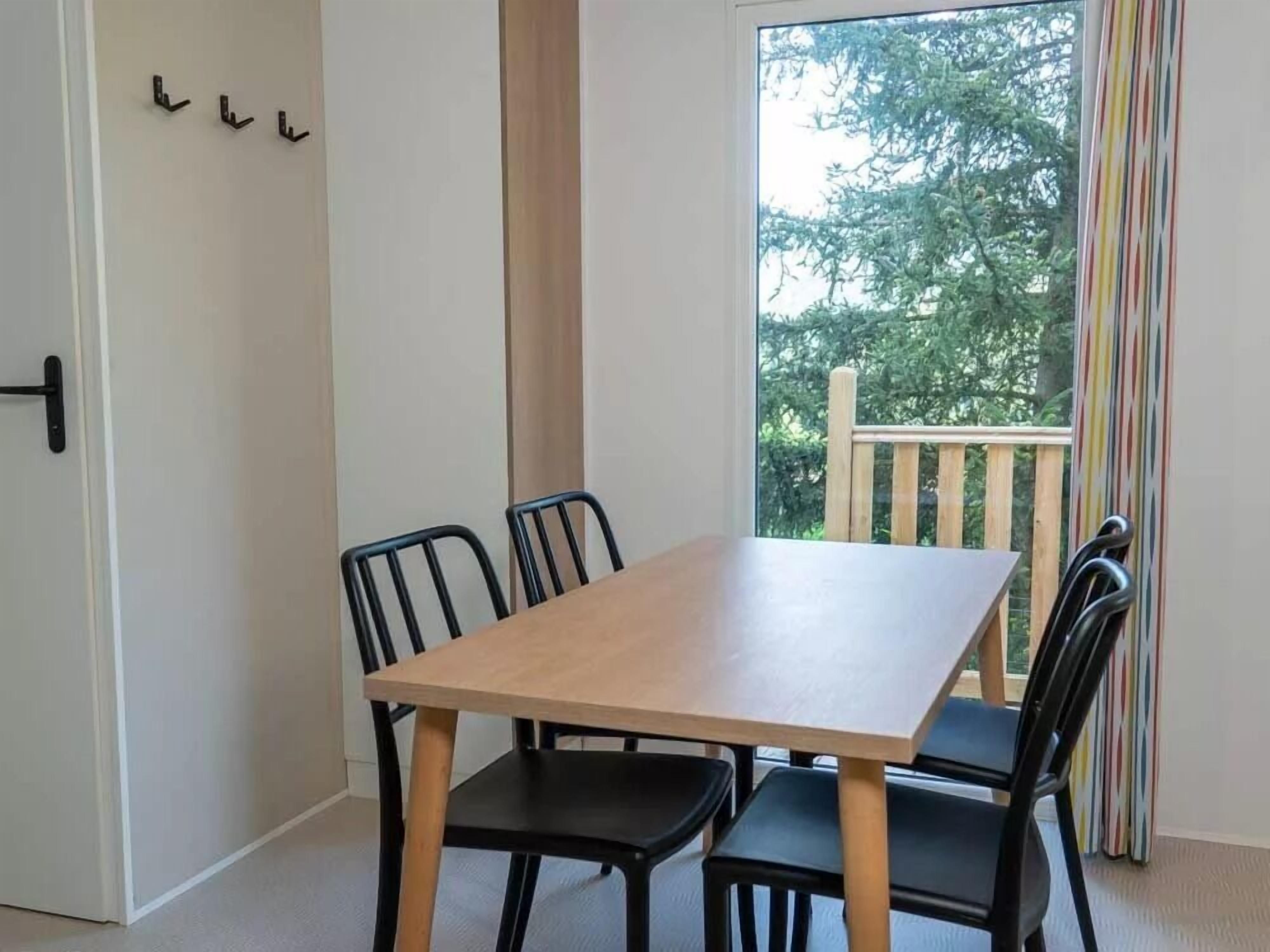 Un comedor con una mesa de madera y cuatro sillas negras. Se observa una puerta con manija negra y tres ganchos negros en la pared. Una ventana ofrece vistas a árboles y a una terraza de madera.