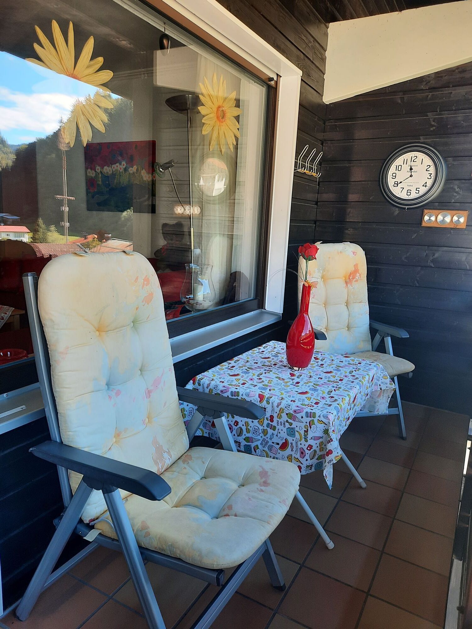 Eine Terrasse mit zwei Sitzgelegenheiten, einem kleinen Tisch mit buntem Tischtuch, einer roten Vase und einer Wanduhr.