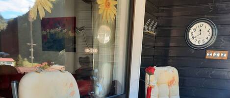 Eine Terrasse mit zwei Sitzgelegenheiten, einem kleinen Tisch mit buntem Tischtuch, einer roten Vase und einer Wanduhr.