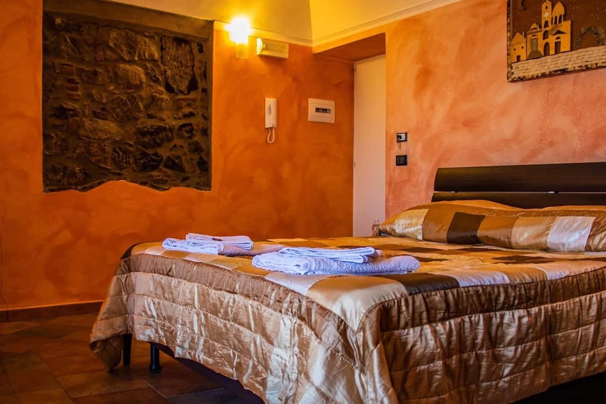 A bedroom with a stone wall, a bed with a brown quilt, and a nightstand with a lamp.