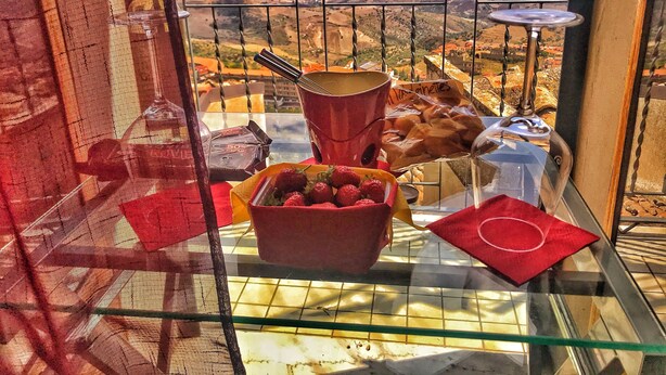 Un balcón con vistas a un paisaje montañoso, una mesa de vidrio con un mantelito rojo, una cesta de fresas y una barra de chocolate.