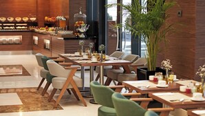 Un comedor moderno con mesas y sillas de madera, un bufé con frutas y bebidas, y una planta grande en maceta.