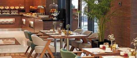 A modern dining area with wooden tables and chairs, a buffet with fruits and beverages, and a large potted plant.