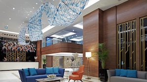 Un vestíbulo moderno con un sofá azul, una silla naranja y un sofá gris. Se observa una lámpara decorativa colgante y un mostrador de recepción al fondo.