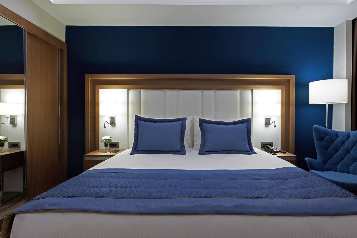 Habitación de hotel con una cama grande, dos lámparas de noche, una mesita de noche, una silla y un espejo.