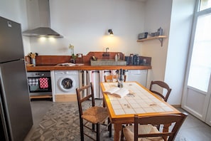 A compact kitchen with a refrigerator, oven, washing machine, and a dining table with chairs. - Charm in the heart of the vineyards (Malras)