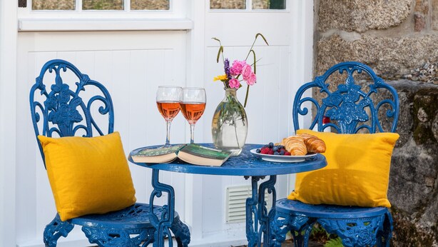 A blue metal table with two chairs, a book, two glasses of red wine, a vase with flowers, and a plate with croissants and berries.