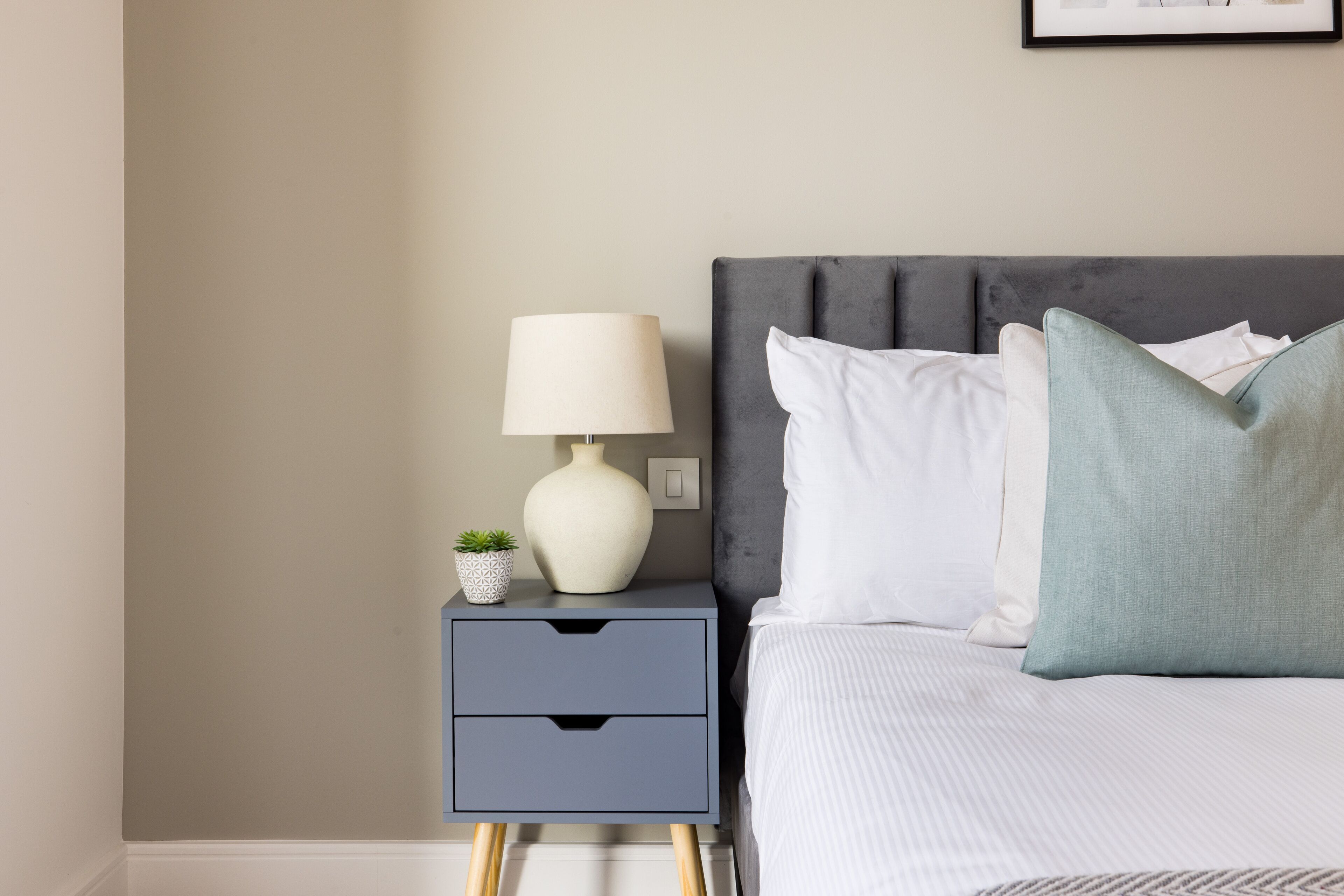 Een netjes opgemaakt bed met witte lakens en een grijs hoofdbord, een nachtkastje met een lamp en een kleine potplant.