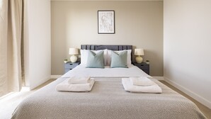 A neatly arranged hotel room with a bed, two bedside tables with lamps, a framed artwork on the wall, and a large window with curtains. - The Wembley Apartment (Wembley)