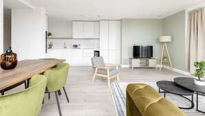 A modern living room with a wooden dining table, green chairs, a grey armchair, a black sofa, a TV, and a floor lamp. - The Wembley Apartment (Wembley)