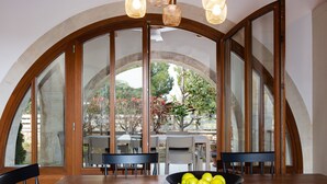 A dining area with a wooden table, chairs, and a bowl of fruit. Large arched windows provide a view of the outdoors.