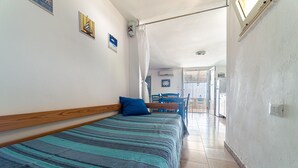 Ein schmaler Flur, der zu einem Schlafzimmer mit Bett, blauem Kopfkissen und Fenster führt.