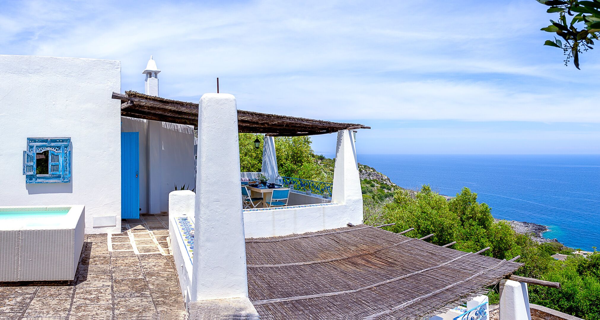 A white building with a blue door and window, a covered outdoor seating area, and a view of the sea.
