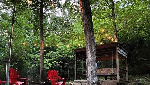 Een terras met rode stoelen en een houten constructie, omgeven door bomen en lichtslingers.