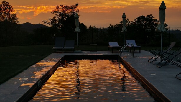 A swimming pool with lounge chairs and umbrellas, surrounded by trees and a sunset sky. - THERESA SULLA COLLINA (Passerano Marmorito)