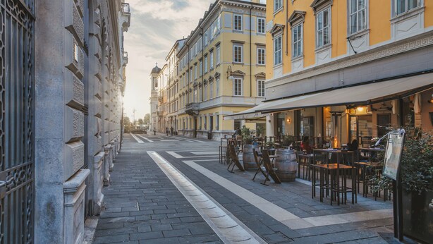 Una calle flanqueada por edificios, con mesas al aire libre en un café, y un cielo despejado.