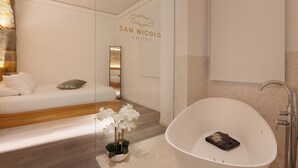 Una habitación de hotel moderna con una bañera blanca, un sofá con cojines, una orquídea en maceta y un espejo montado en la pared.