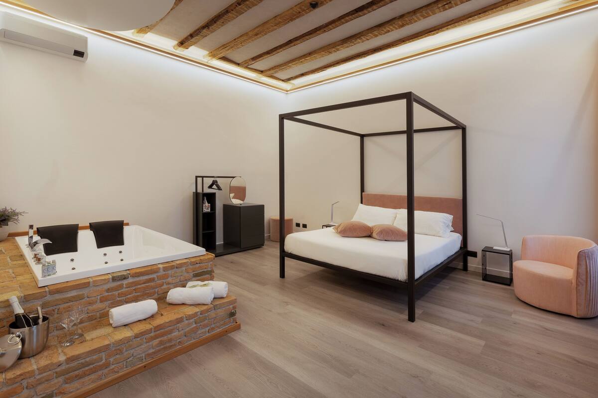 A modern bedroom with a four-poster bed, a built-in bathtub, and a seating area with a cushioned chair.