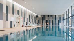 A modern indoor swimming pool with clear glass walls, a tiled floor, and a row of white lounge chairs along the side. - Diamant Hotel y Aparthotel (Capdepera)