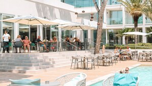 An outdoor pool area with lounge chairs and umbrellas, surrounded by a modern building with large windows. - Diamant Hotel y Aparthotel (Capdepera)