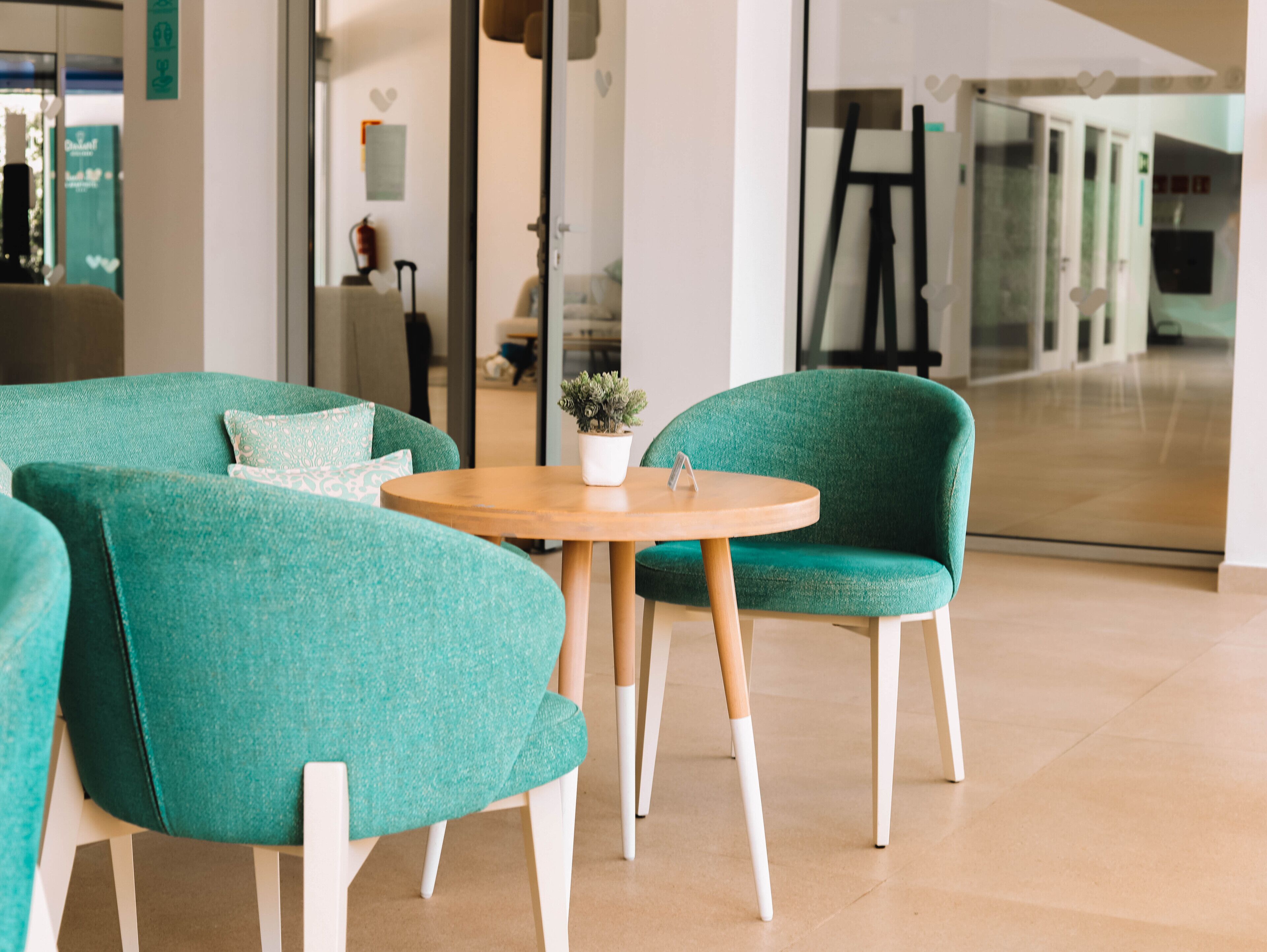 A modern interior with teal upholstered chairs around a wooden table, a small potted plant, and a glass partition reflecting the room.