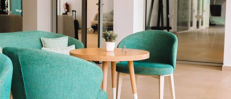 A modern interior with teal upholstered chairs around a wooden table, a small potted plant, and a glass partition reflecting the room.