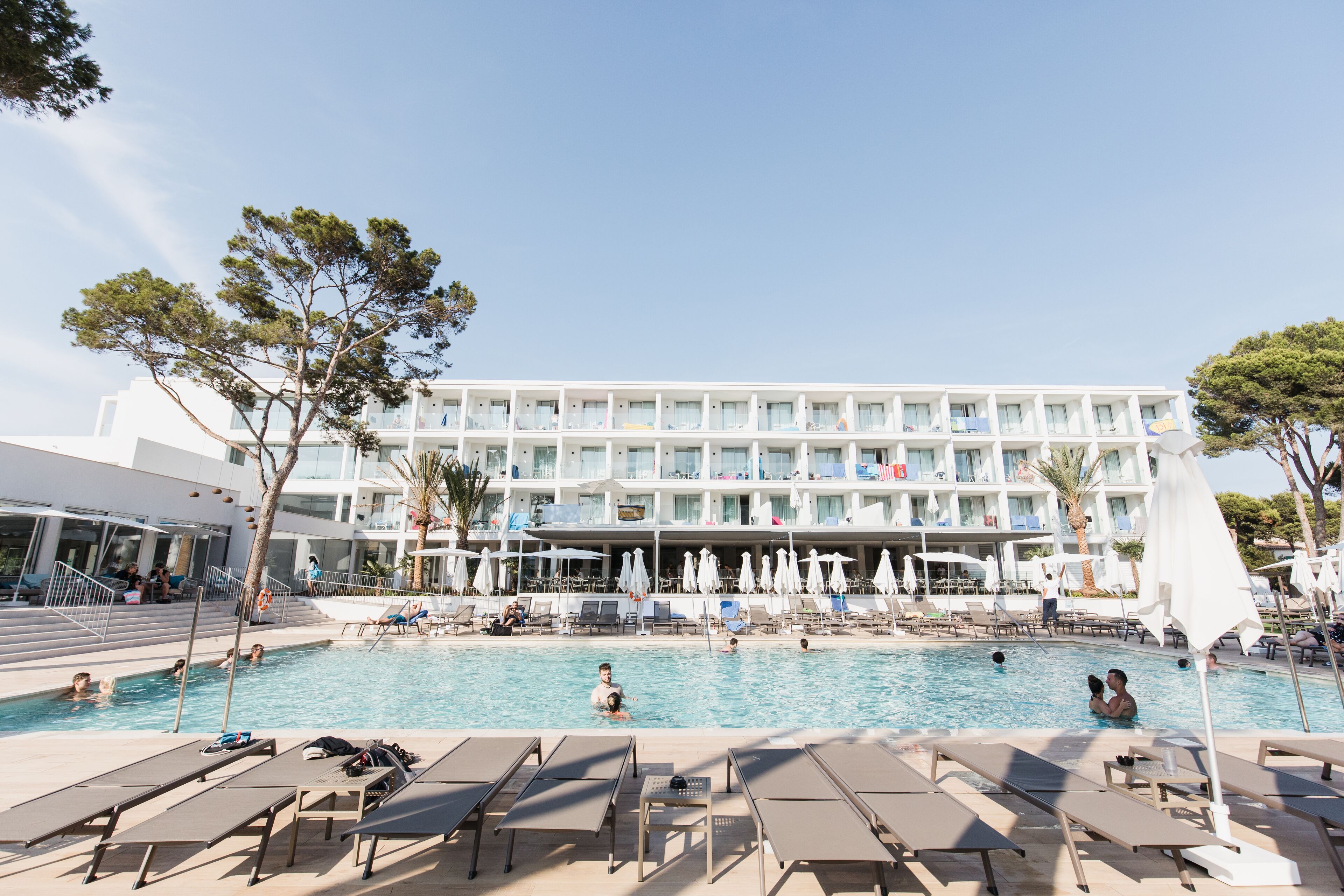 A hotel pool area with sun loungers, umbrellas, and a building with balconies.