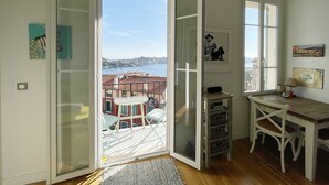 Une chambre avec un balcon, un bureau, une chaise et une vue sur les bâtiments et l’eau.