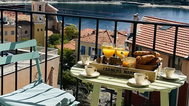 Un balcon avec une table dressée pour le petit-déjeuner, offrant une vue panoramique sur la côte, avec des bâtiments et une colline en arrière-plan.