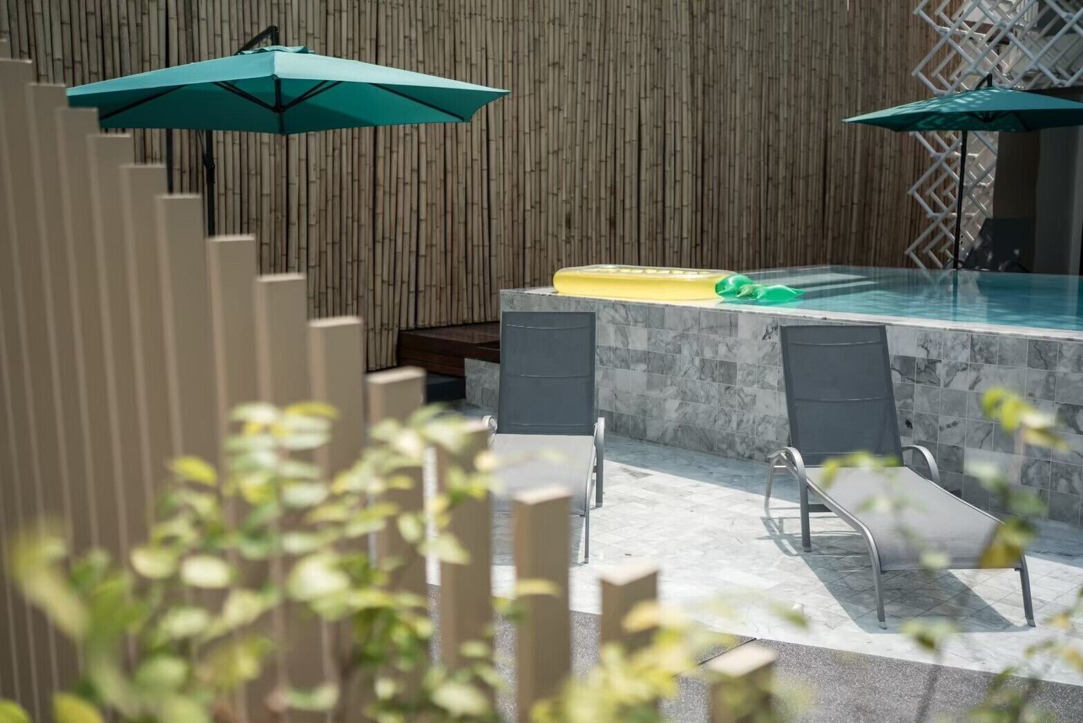 Zona de piscina exterior con valla de madera, dos sillones de descanso y una plataforma flotante amarilla.