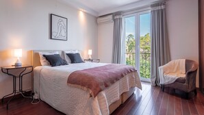 Un dormitorio con una cama grande, dos mesitas de noche, una silla y un ventanal con cortinas.