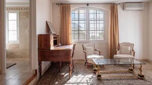 Una habitación con un escritorio de madera, dos sillas, una mesa de centro de vidrio y un ventanal con cortinas.