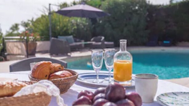 Desayuno con croissants, ciruelas, un vaso de jugo de naranja y una botella de jugo de naranja junto a una piscina.