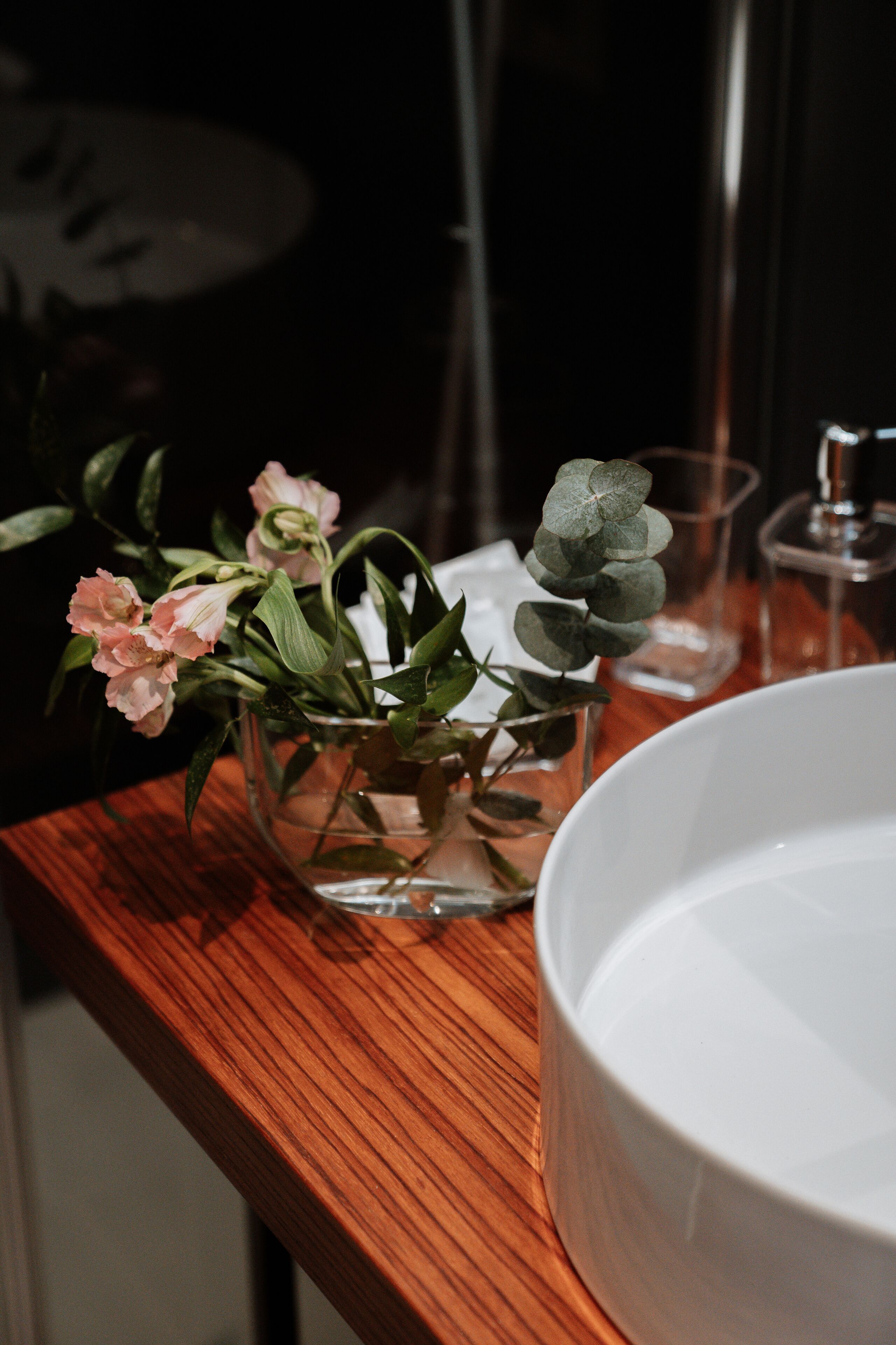 Ein Holztisch mit einer Glasvase, die rosafarbene Blumen und grüne Blätter enthält, ein klares Glas und eine weiße Keramikschale.