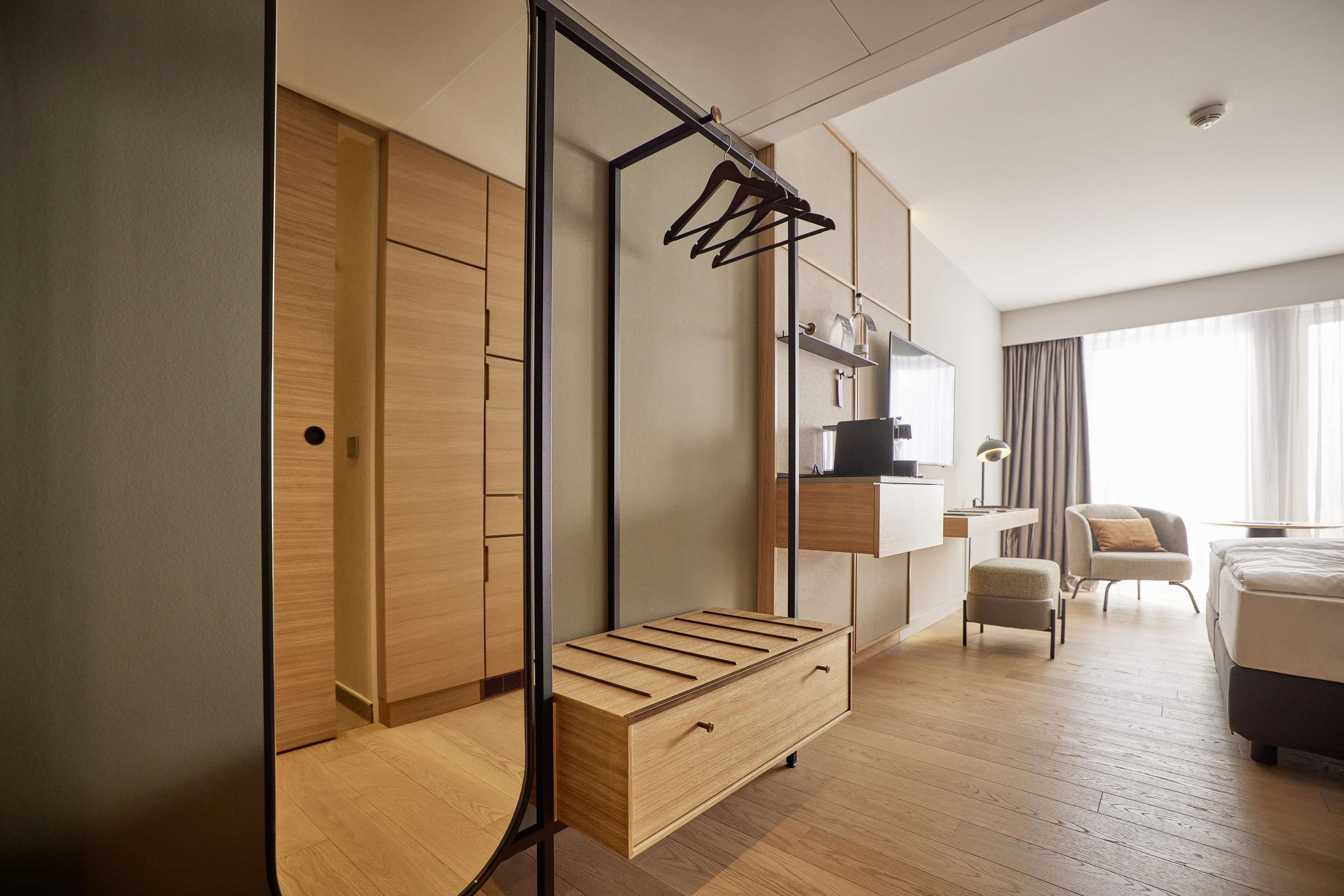 A modern hotel room with a wooden wardrobe, a built-in bench, a desk with a chair, and a bed.