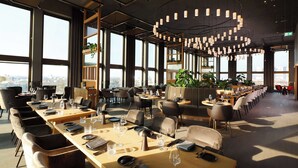 A modern restaurant with long communal tables, individual seating, and a view of the city through large windows.