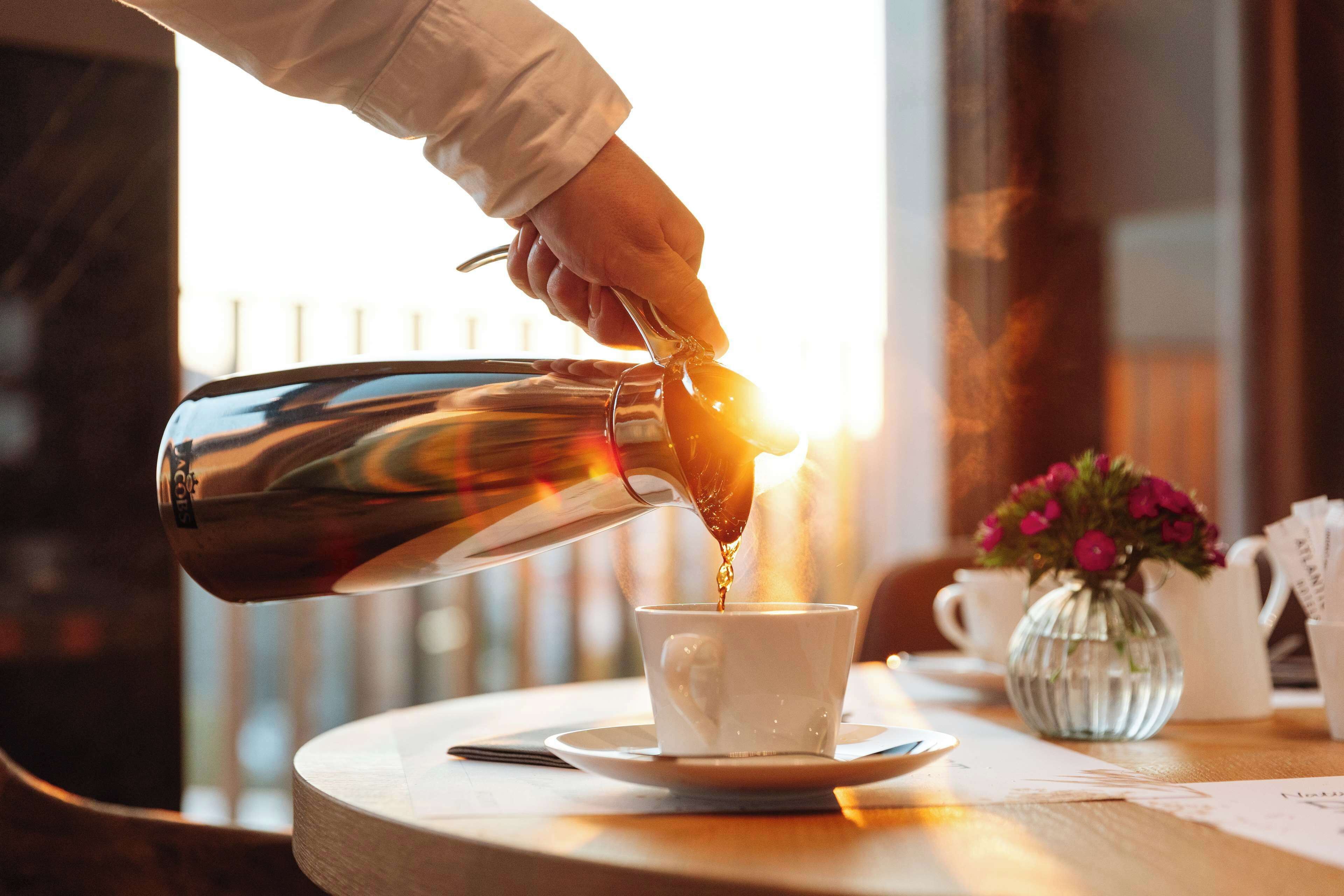 A hand pouring coffee into a cup on a table with a vase of flowers and a menu.