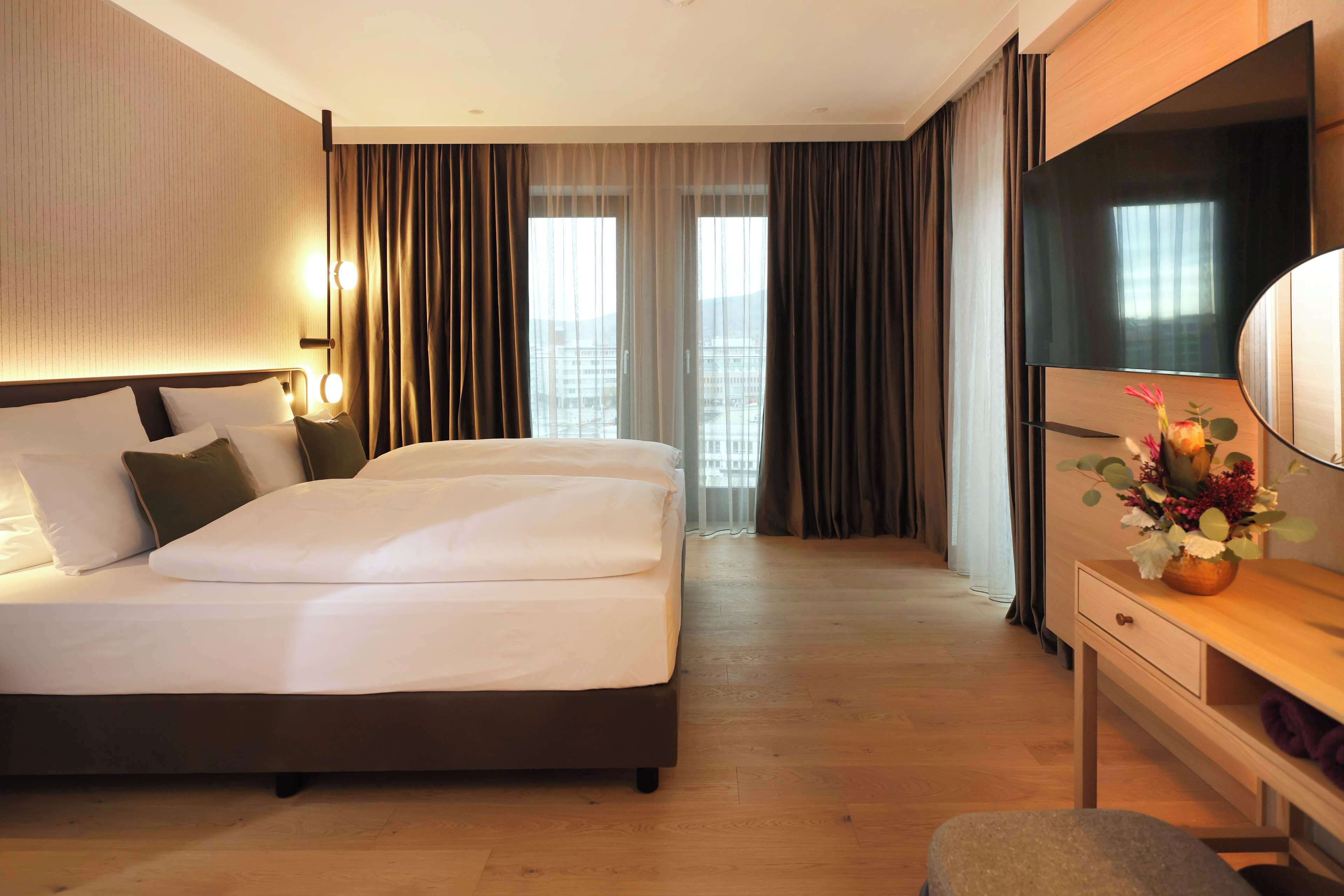 Habitación de hotel con una cama grande, un escritorio con televisor y vistas a la ciudad desde las ventanas.